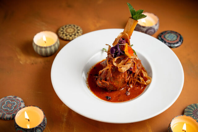 Spiced lamb shank curry garnished with edible flowers and mint, surrounded by glowing candles on a warm-toned table.